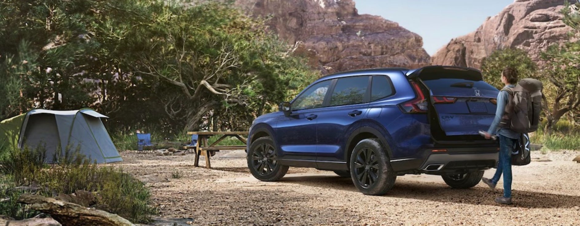 A white person with a backpack using the power tailgate of a blue 2026 Honda CR-V SUV that is parked near a campsite with mountains in the background