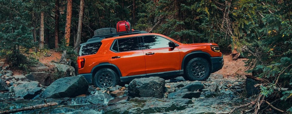 An orange 2026 Honda Passport with a loaded roof rack crossing a rocky river