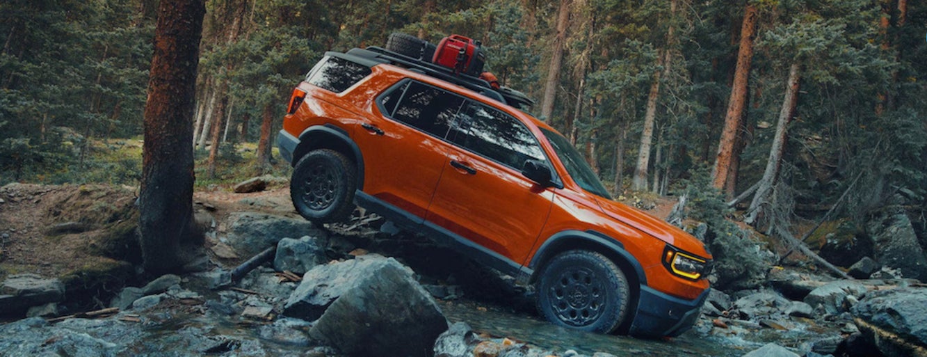 An orange 2026 Honda Passport SUV driving into a rocky riverbed in a forest