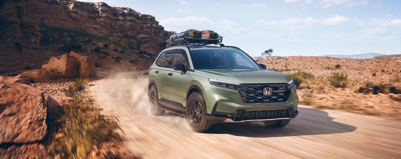 A green 2026 Honda CR-V SUV driving down a dirt road through the desert, with a mesa in the background