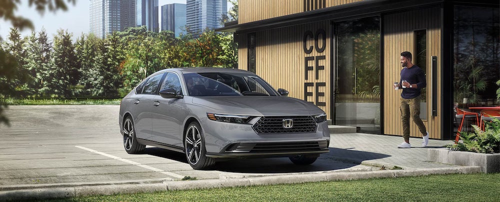 A gray 2025 Honda Accord sedan parked in front of a coffee shop, a man with brown skin holding coffees walking toward the car